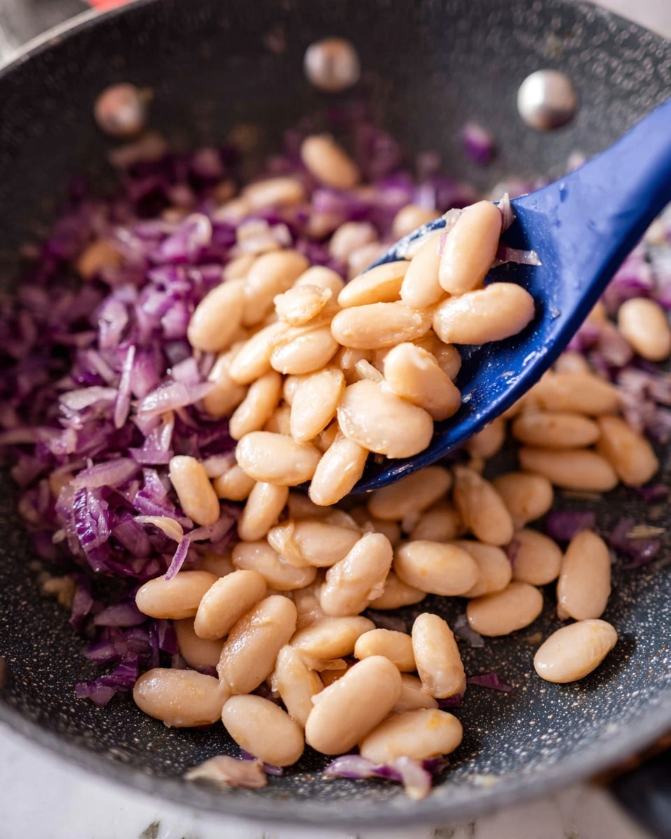 The image shows a close-up of a dark gray speckled pan with cooked white beans layered on top of a base of finely chopped cooked purple onions. In the center, a blue spoon lifts several beans, highlighting their smooth, light beige texture. The pan’s metal rivets and a white marbled surface are faintly visible around the edges. Photo taken with an iphone --ar 4:5 --v 7