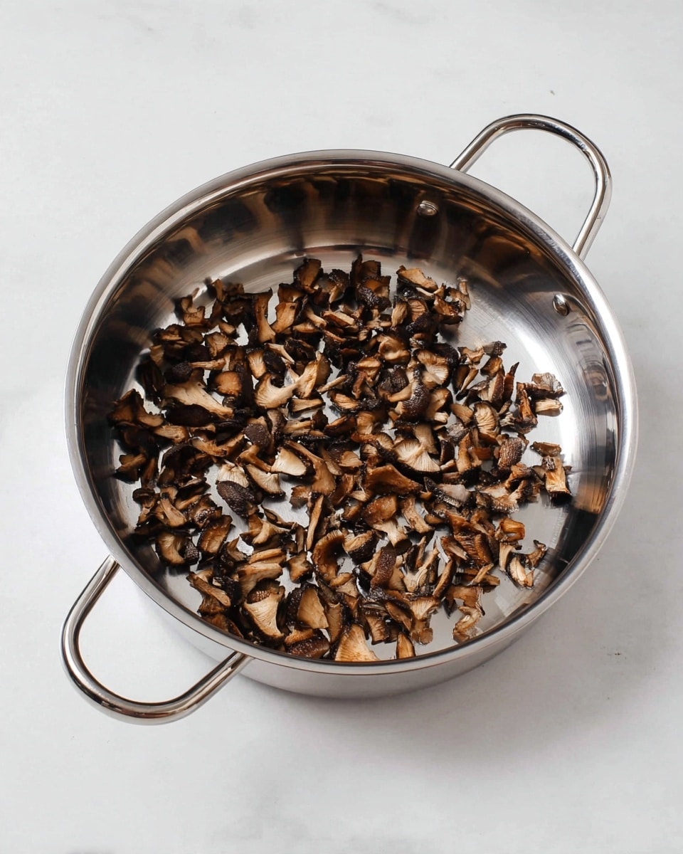 A shiny stainless steel pan with two handles is shown on a white marbled surface, filled with a single layer of small, cooked mushroom pieces. The mushrooms are a mix of dark brown and light brown shades, with some pieces showing a slightly crisp, caramelized texture. The pan reflects light, adding a glossy look to the metal, while the mushrooms are scattered unevenly across the bottom. The overall image has a clean and simple feel. photo taken with an iphone --ar 4:5 --v 7