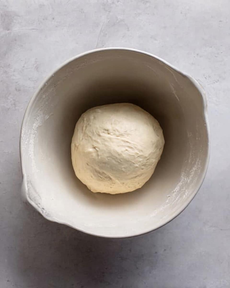 A single round ball of dough with a smooth, slightly shiny surface sits in the center of a large white mixing bowl with a handle. The dough is off-white and looks soft and elastic. The bowl rests on a white marbled surface, giving a clean and bright background that contrasts with the dough and bowl. photo taken with an iphone --ar 4:5 --v 7