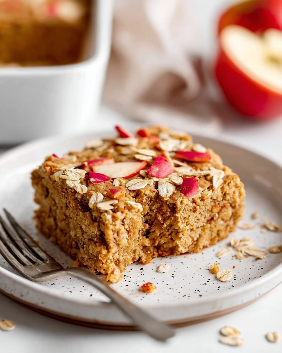 A thick square piece of baked oat cake sits on a white plate with small dark specks; the cake is light brown and textured with visible oats and small apple pieces inside and on top, with scattered red apple skin bits and oat flakes decorating the surface. The plate is on a white marbled background with a silver fork resting beside it, and part of the cake has a bite taken out, showing its soft, moist interior. In the blurry background, another slice of cake is partially visible in a white bowl, along with a blurred red apple half, photo taken with an iphone --ar 4:5 --v 7
