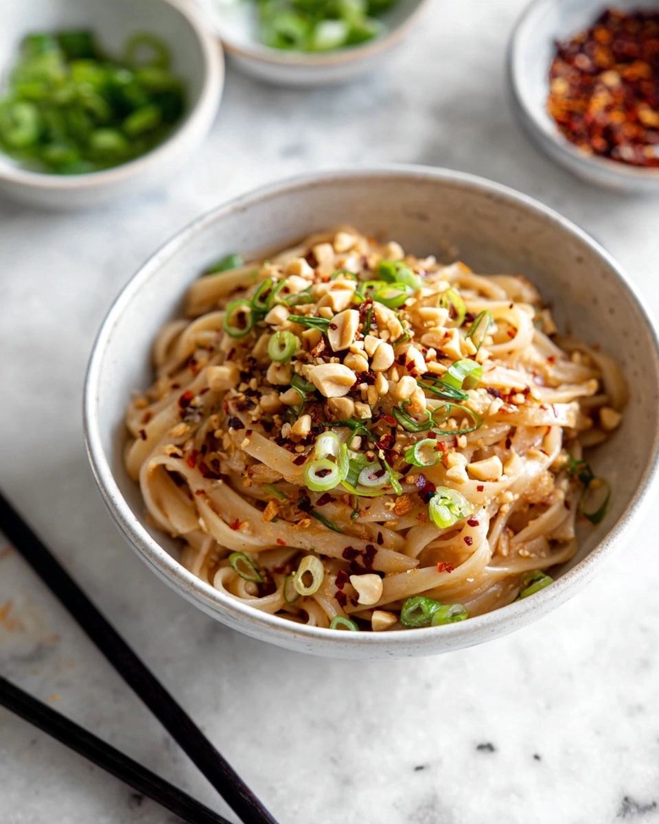 A white bowl full of thick, light brown noodles covered with sliced green onions and chopped peanuts scattered on top, with red chili flakes sprinkled throughout. The noodles look soft and slightly shiny, mixed well with the toppings. The bowl rests on a white marbled surface, with a pair of black chopsticks nearby. In the background, small white bowls hold more green onions and chili flakes, slightly out of focus. photo taken with an iphone --ar 4:5 --v 7
