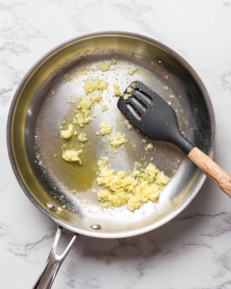 A shiny silver frying pan with light yellow minced garlic frying in a small pool of clear oil, positioned on a white marbled surface, a black spatula with a wooden handle rests on the right side inside the pan, showing some garlic stuck to it. photo taken with an iphone --ar 4:5 --v 7