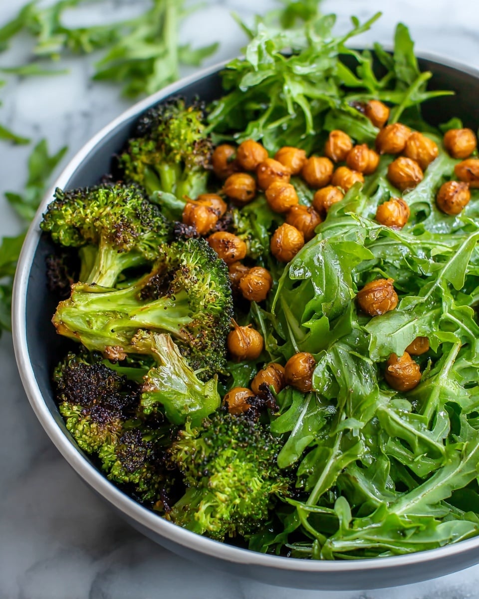 A close-up view of a salad in a white bowl, showing three layers: at the bottom, pieces of roasted broccoli with browned edges and uneven char; the middle layer is bright green fresh arugula leaves with a slightly wet and shiny look; the top layer has small, round, golden roasted chickpeas scattered across the arugula. The bowl rests on a white marbled surface, with some scattered green leaves in the background. Photo taken with an iphone --ar 4:5 --v 7
