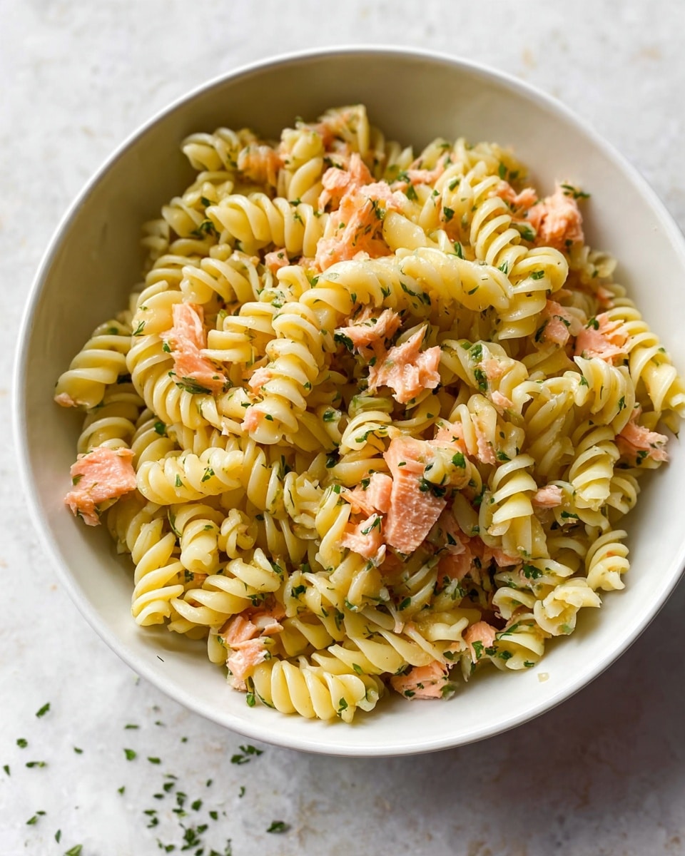 A white bowl filled with spiral pasta, light yellow in color, mixed evenly with small pieces of pink salmon scattered throughout. There are tiny green herb bits sprinkled on top and mixed in, adding fresh color contrast. The bowl sits on a white marbled surface with a few herb bits around it. The texture of the pasta is smooth and slightly shiny, and the salmon pieces look soft and flaky. Photo taken with an iphone --ar 4:5 --v 7